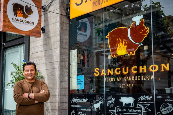 A man poses for a portrait. He is on the left and on the right is the window of the restaurant with the logo